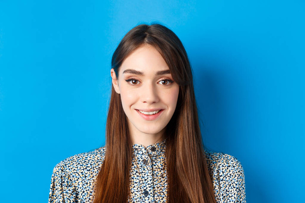 Close-up portret van aantrekkelijke natuurlijke meisje met lang haar en make-up, glimlachen gelukkig en vriendelijk op de camera, blauwe achtergrond - Foto, afbeelding