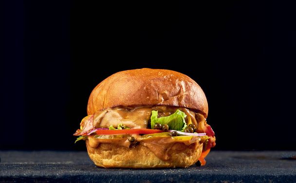 Ζουμερό burger με μοσχάρι, τυρί, καραμελωμένα κρεμμύδια και ντομάτα, σάλτσα σε σκούρο φόντο. - Φωτογραφία, εικόνα