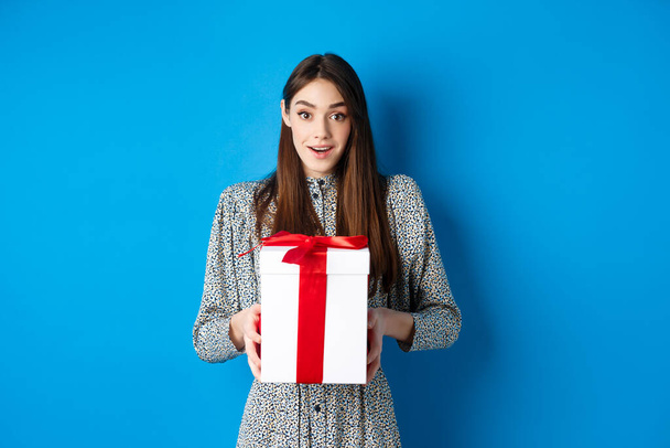 Valentines day concept. Surprised happy girl receiving romantic gift and looking thankful at camera, standing on blue background - Fotó, kép
