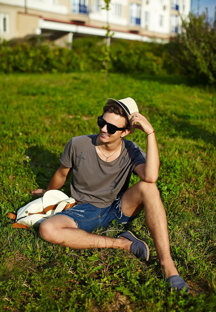 Portrait of young attractive modern stylish man in casual cloth in hat in glasses sitting in the park in green grass - Foto, afbeelding