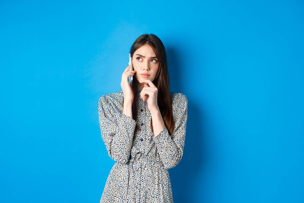 Страшная женщина разговаривает по телефону и думает, глядя в сторону вдумчивый, стоя на синем фоне - Фото, изображение