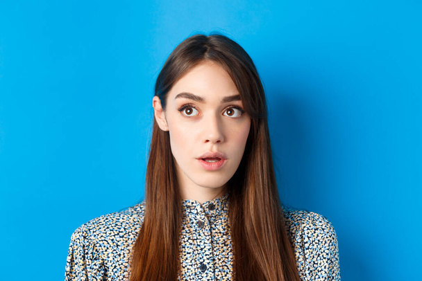 Κοντινό πλάνο πορτρέτο της έκπληκτης νεαρής γυναίκας με φυσική ομορφιά, κοιτάζοντας την πάνω αριστερή γωνία και λαχανιάζοντας, στέκεται σε μπλε φόντο - Φωτογραφία, εικόνα