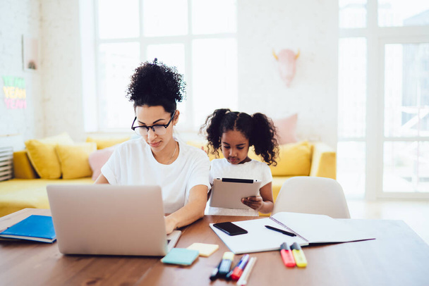 African American little girl standing in bright room and browsing tablet while mother in glasses working and typing on laptop - Zdjęcie, obraz