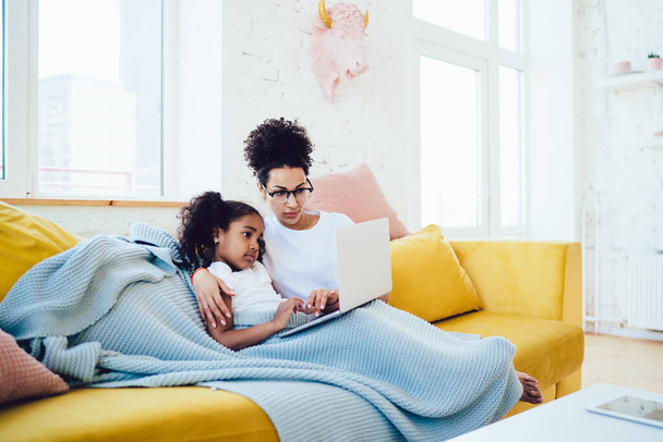 Ethnic mother in glasses sitting with cute child on yellow sofa under blue blanket while watching video in social media on laptop - Φωτογραφία, εικόνα