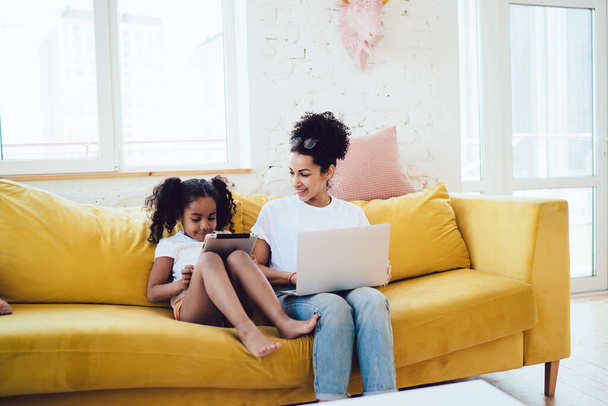 African American mother in white shirt and blue jeans with eyeglasses on head looking at daughters with tablet while sitting on yellow sofa together - Photo, image