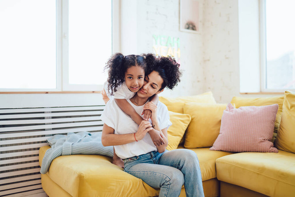 Loving African American woman and charming girl in casual clothes hugging and sitting on cozy sofa in modern light room and looking at camera - 写真・画像