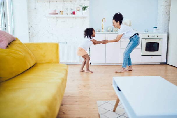 Happy African American mother in casual clothing with curly hair playing with smiling daughter while standing in modern kitchen of studio apartment - Foto, Imagen