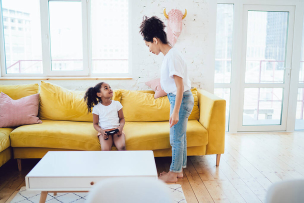 Ethnic mother in casual clothes standing in bright room and looking at daughter while adorable girl with controller in hand smiling - 写真・画像