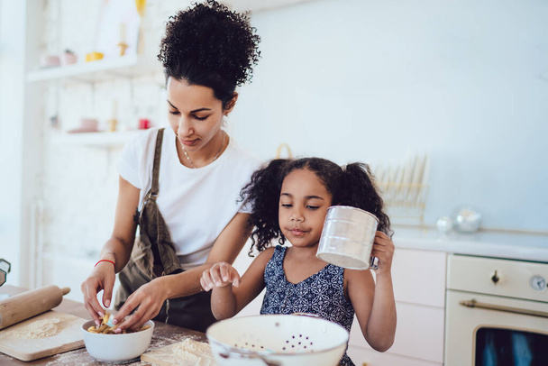 Young ethnic curly woman in causal outfit standing with little daughter holding mug at table preparing breakfast in kitchen at home looking at camera  - Фото, изображение