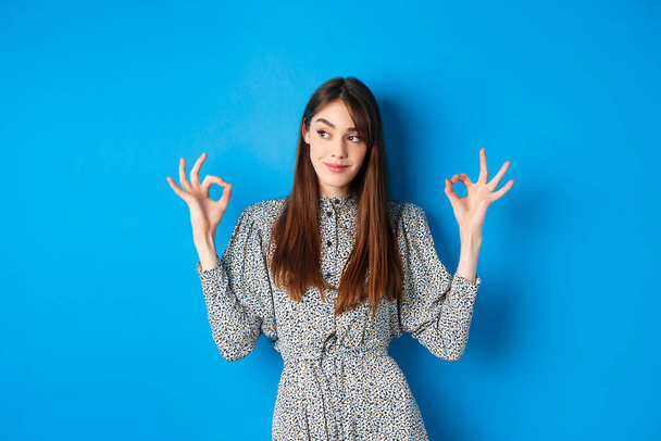 Not bad. Pleased smiling woman with long hair, wearing dress, showing okay signs and looking satisfied, praising good job, looking left at logo, blue background - Fotografie, Obrázek