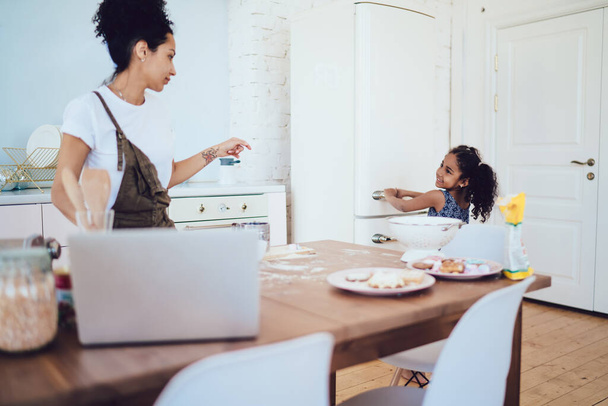 African American mother telling smiling daughter with curly hair to open fridge and take ingredients while cooking in kitchen of modern studio apartment  - Foto, Imagem