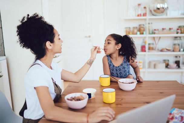 Beautiful young mother feeding cute daughter with cereal while both sitting at table and eating breakfast in kitchen at home - Foto, Imagem