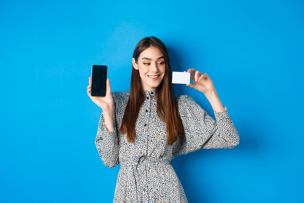 Online shopping. Beautiful female model showing empty cellphone screen and plastic credit card, smiling pleased, buying in internet store, blue background - Photo, Image