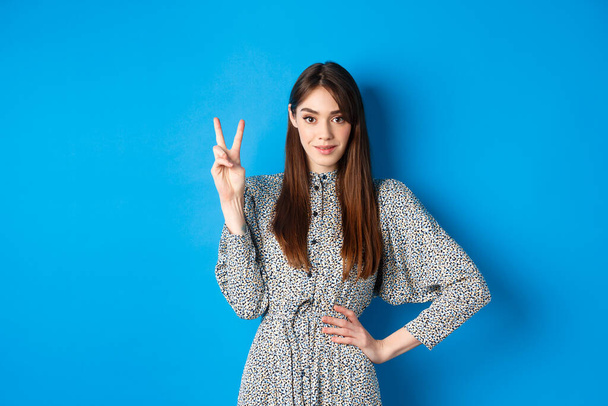 Young beautiful girl in dress with natural long hair, showing number two with fingers and smiling, standing against blue background - 写真・画像