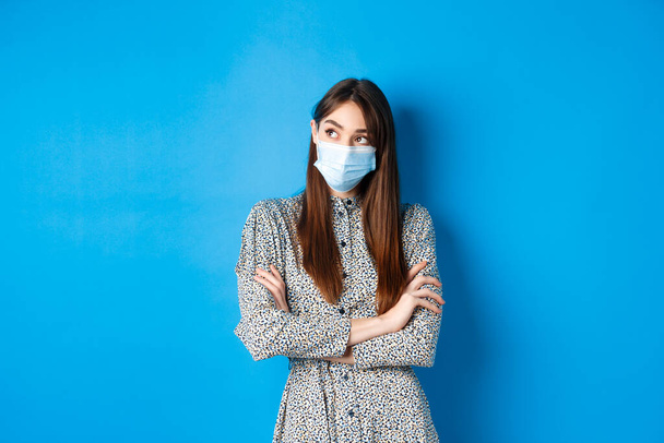 Covid-19, pandemic lifestyle concept. Pensive girl in medical mask look at upper left corner logo, thinking ways of protecting health from coronavirus, blue background - Fotografie, Obrázek