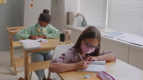 Sklopený záběr dívek v obličejových maskách sedících na lavicích ve třídě a zapisujících do sešitů během výuky na střední škole - Záběry, video