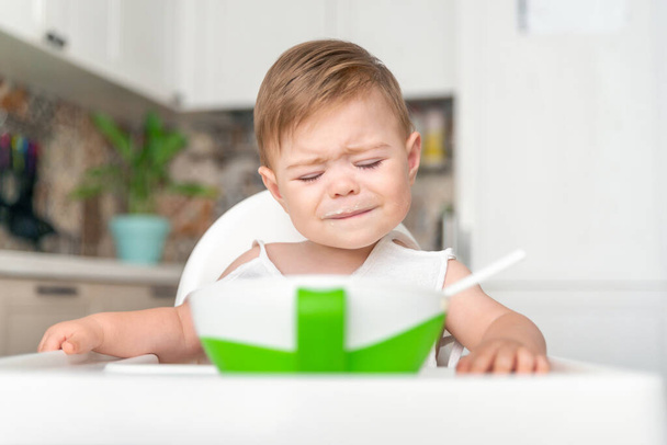 高椅子に座って汚い乱雑な顔をした不幸な小さな白人の子供の少年.赤ん坊の泣き声、気まぐれな、食べることを拒否する - 写真・画像