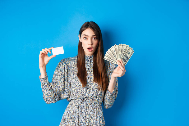 Ενθουσιασμένη τυχερή κοπέλα που δείχνει πλαστική πιστωτική κάρτα και χρήματα δολάριο, λαχανιάσει κατάπληκτος, αγοράζοντας κάτι ακριβό, στέκεται σε μπλε φόντο - Φωτογραφία, εικόνα