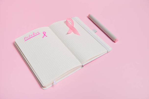 Lettering 1 st October on a diary and a pink ribbon on an empty blank paper sheet, isolated on pink background with copy space. Breast Cancer Awareness Month - Photo, image