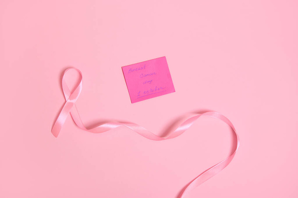 Plocha ležela z dlouhé růžové stuhy s nekonečným koncem a papírové poznámky s nápisem 1. října Den povědomí o rakovině prsu, izolované na růžovém pozadí s prostorem pro text - Fotografie, Obrázek