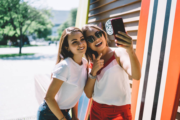 Cheerful young friends in vivid casual clothes taking selfie with smartphone while standing on street against striped wall and vibrant surf boards in sunny summer day - Фото, зображення