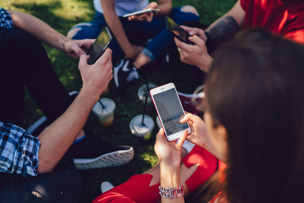 Groep jonge vrienden surfen op mobiele telefoons zitten rond op gras met koppen koffie in de stad in de zomer - Foto, afbeelding