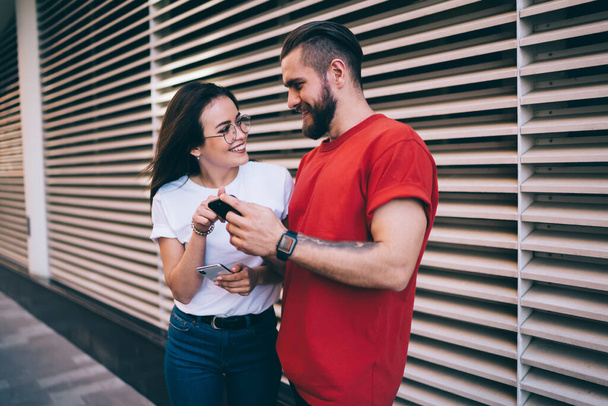 Attractive bearded man and young woman smiling and looking at each other while standing near modern building on city street and using smartphones - 写真・画像