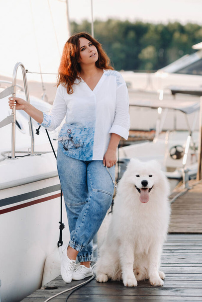 a happy woman with a big white dog stands on the pier near the yacht. - Zdjęcie, obraz