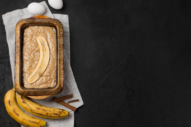 Raw banana bread dough in rectangular baking dish with ingredients on black background - Photo, Image