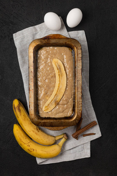 Raw banana bread dough in rectangular baking dish with ingredients on black background - Foto, immagini