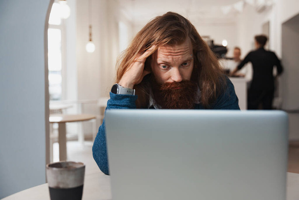 Tired man with red, long hair works at a laptop - Foto, imagen