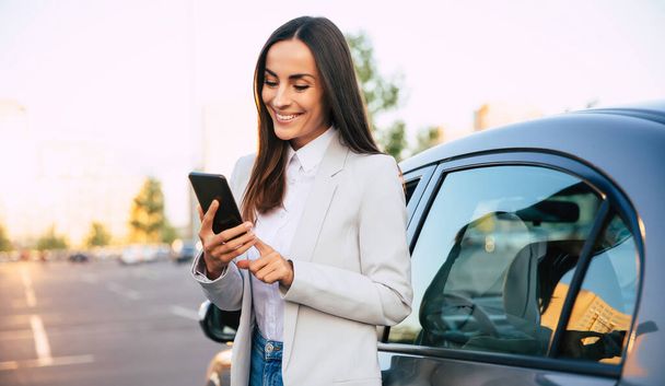 Başarılı bir şekilde gülümseyen, şık giyinmiş çekici bir kadın modern arabanın yanında dururken akıllı telefonunu kullanıyor. - Fotoğraf, Görsel