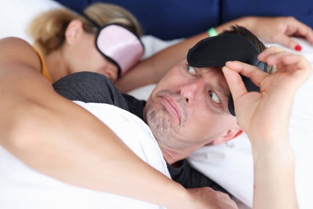 Έκπληκτος άντρας σηκώνει τη μάσκα του ύπνου και κοιτάζει τη γυναίκα να τον αγκαλιάζει στην κρεβατοκάμαρα. - Φωτογραφία, εικόνα