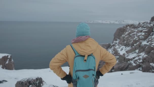 Un homme en vêtements brillants et avec un sac à dos se dresse au sommet d'une falaise enneigée et admire la mer. Concept de solitude et de réflexion - Séquence, vidéo