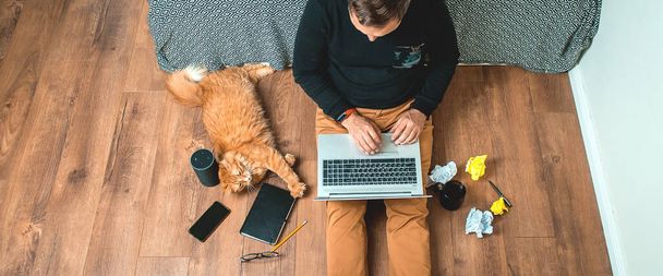 Work from home on creative workspace. Man sitting on floor with laptop computer and smart speaker. Ginger pet cat lying. Home office. Remote work - Photo, Image