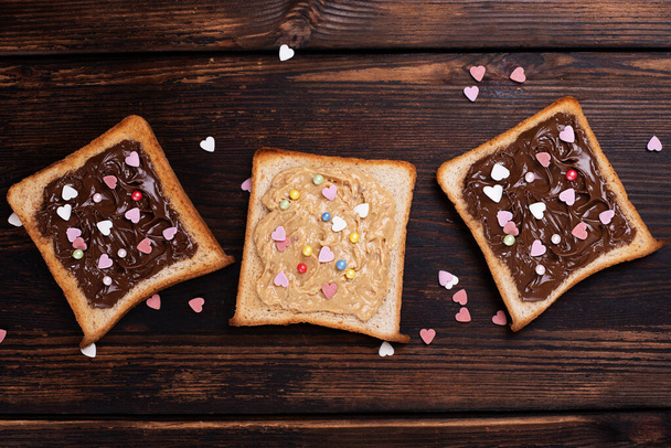 Three toast with chocolate paste and peanut butter sprinkled with colorful hearts on a dark wooden background, top view. - Foto, afbeelding