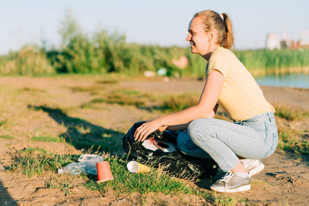 Fiatal női önkéntes elégedett szedés szemetet, műanyag palackok és kávéscsészék, tiszta strand egy tengerrel. A nő szemetet gyűjt. A környezetszennyezés fogalma. Föld napja - Fotó, kép
