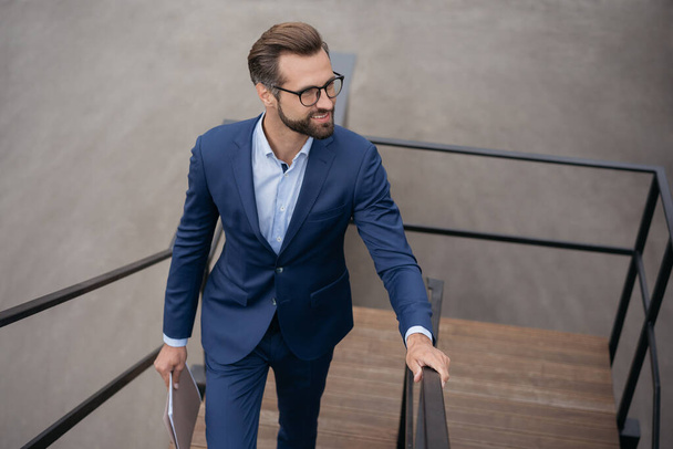 Young confident businessman holding financial documents walking up stairs looking away at copy space. Successful business, career concept - Photo, Image