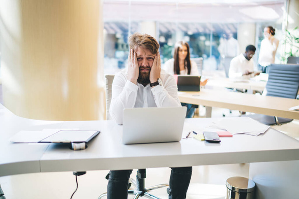 Portrait d'un employé masculin malheureux assis à la table de bureau avec ordinateur portable et se sentant mal à la tête et le stress des heures supplémentaires à l'intérieur du bureau, freelance épuisé fatigué du travail à distance - Photo, image