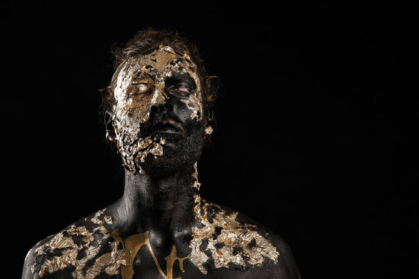 Handsome young man with black and golden paint on body against dark background - Foto, Imagem