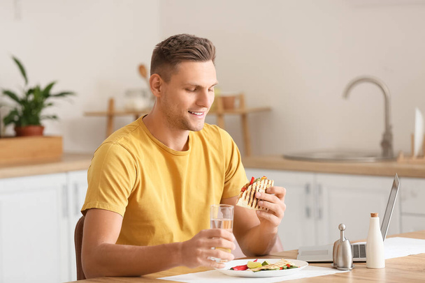 Knappe jongeman met laptop lekker quesadilla eten thuis - Foto, afbeelding