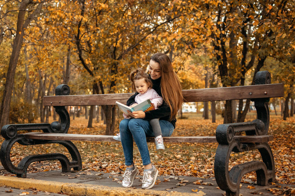 Nuori nainen lapsenvahti ja lapsi vauva tyttö lukea kirjaa syksyllä puistossa. Onnellinen perhe äiti ja lapsi ulkona syksyllä puistossa - Valokuva, kuva