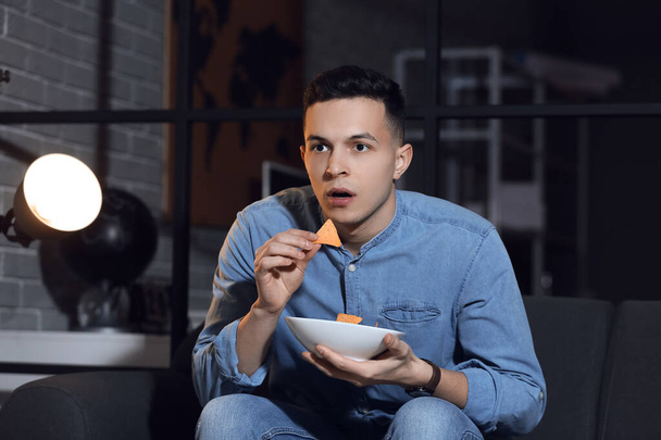 Komea nuori mies syö maukkaita nachoja katsoessaan televisiota kotona myöhään illalla - Valokuva, kuva