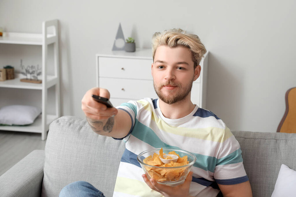 Handsome young man eating tasty nachos while watching TV at home - Photo, Image