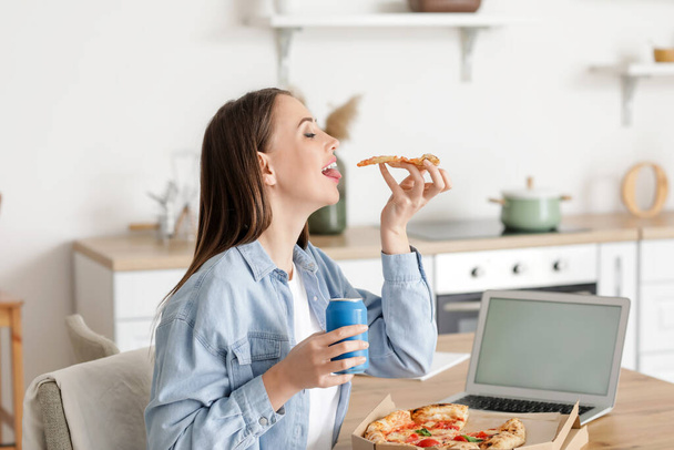 Beautiful young woman with laptop eating tasty pizza in kitchen - Photo, image