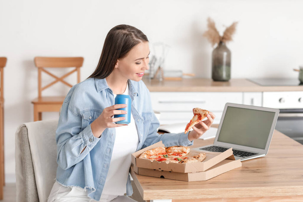 Beautiful young woman with laptop eating tasty pizza in kitchen - Fotografie, Obrázek