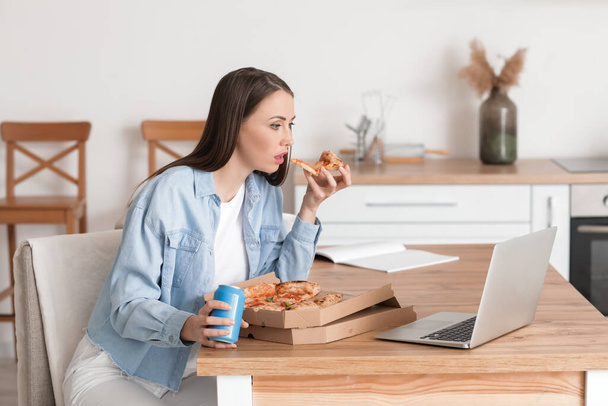 Beautiful young woman with laptop eating tasty pizza in kitchen - Fotó, kép