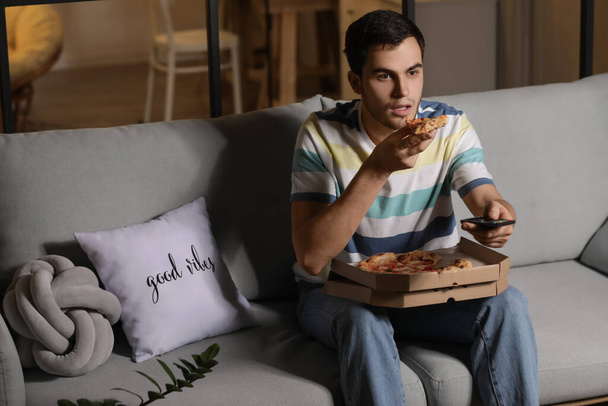 Handsome young man eating tasty pizza while watching TV at home in evening - Valokuva, kuva