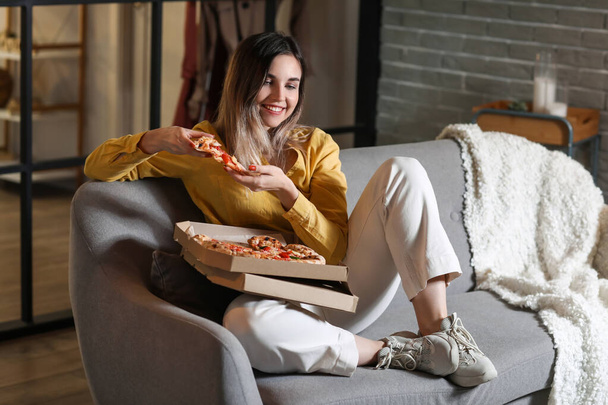 Beautiful young woman eating tasty pizza at home in evening - Fotó, kép