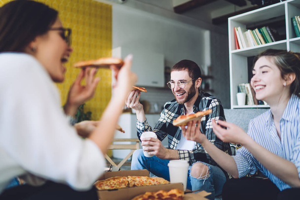 Young man and woman laughing and eating delicious pizza while sitting near blurred female friend during party in cozy living room - Zdjęcie, obraz